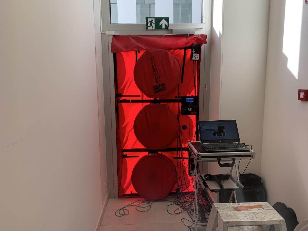Blowerdoor testopstelling in een deuropening met rode meetdoek en ventilatoren, aangesloten op meetapparatuur en laptop voor luchtdichtheidsmeting van een gebouw.