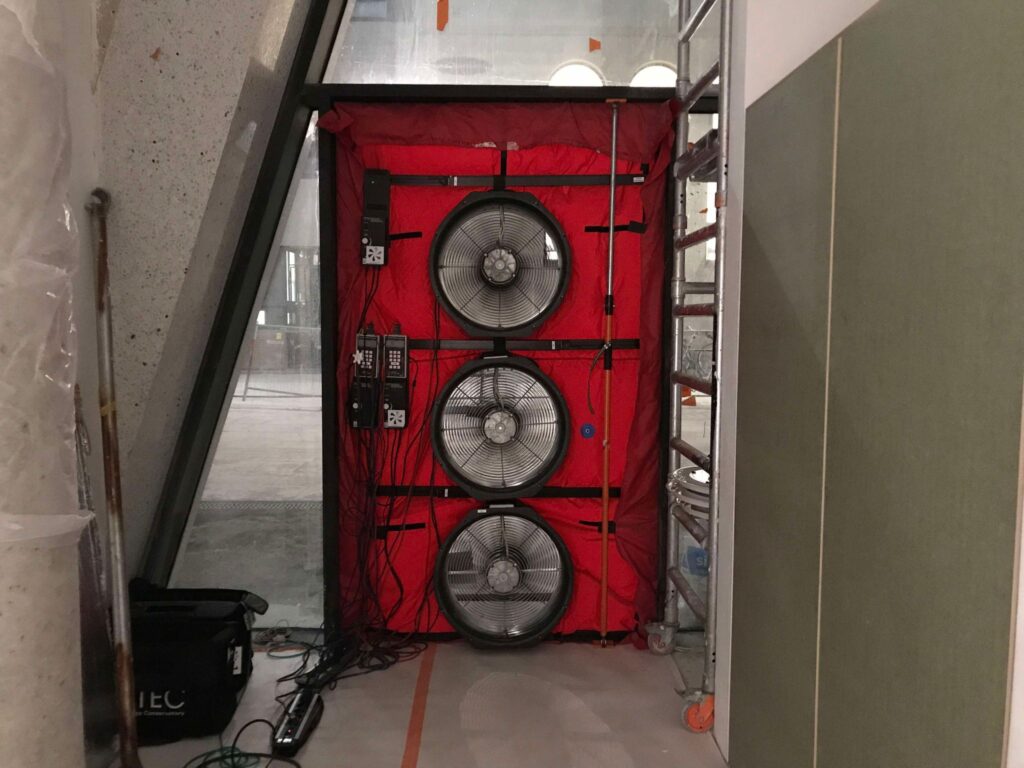 Blowerdoor testopstelling met drie ventilatoren in een deuropening, geplaatst in een bouwomgeving voor het meten van de luchtdichtheid van een gebouw.