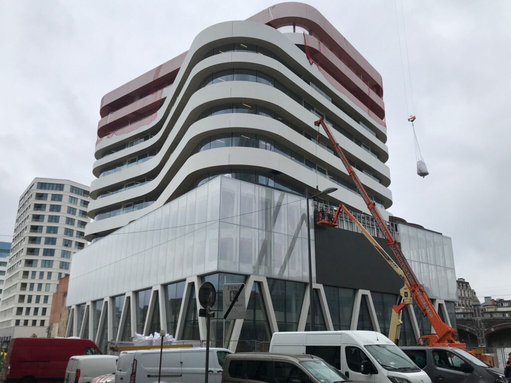 Modern, golvend vormgegeven gebouw met meerdere verdiepingen en glazen plint, gefotografeerd tijdens de bouwfase met hijskraan en werkzaamheden zichtbaar.