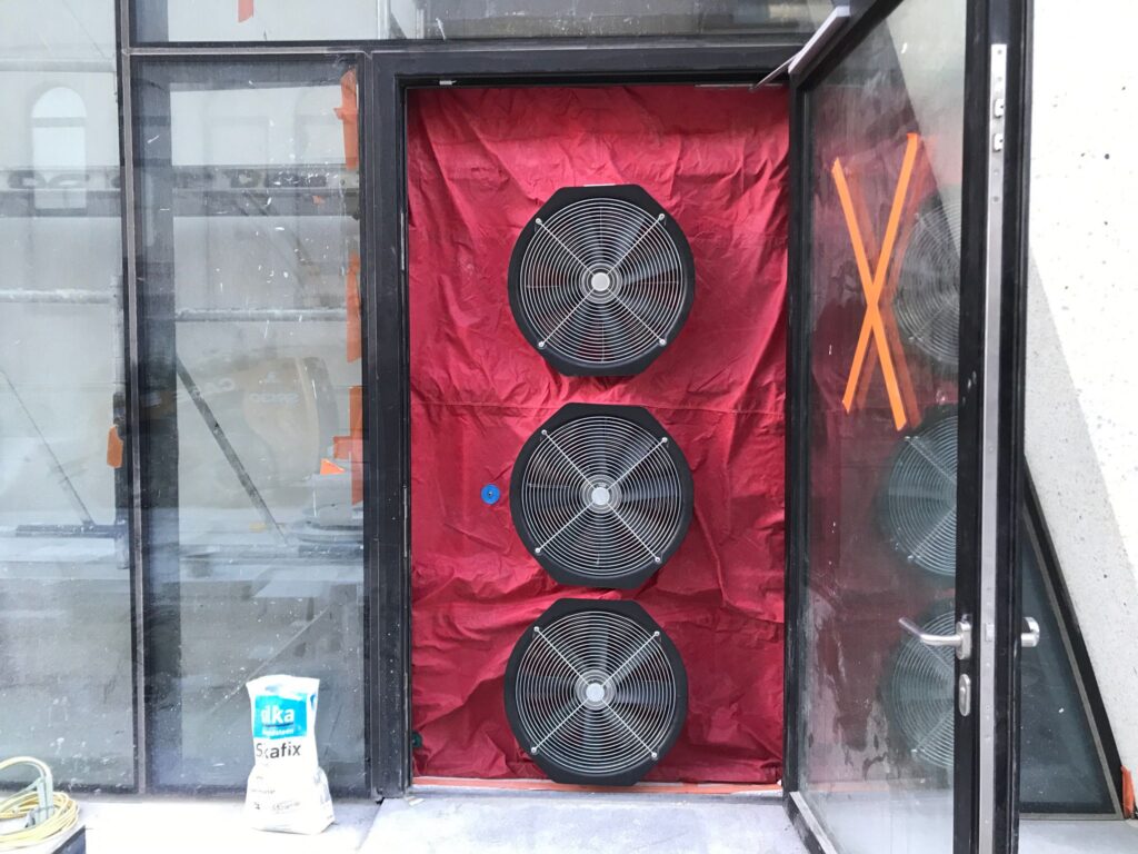 Blowerdoor testopstelling met drie ventilatoren in een glazen deuropening, geplaatst in een gebouw in aanbouw voor luchtdichtheidsmeting.