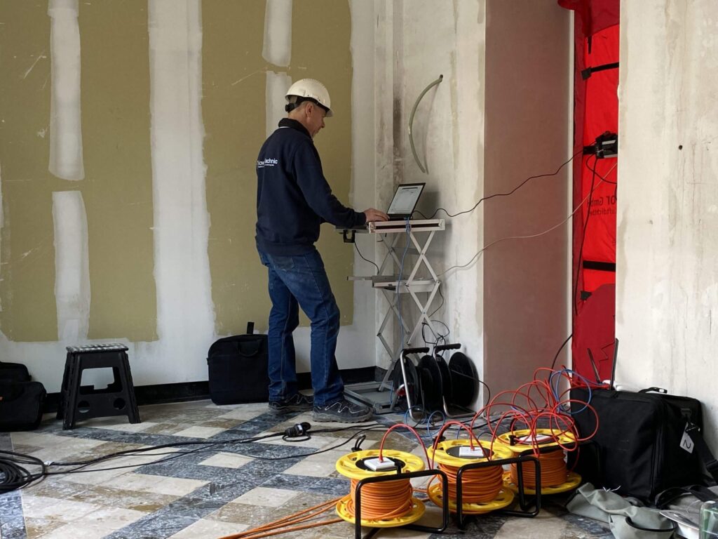 Technicus voert een blowerdoortest uit in een gebouw in afwerking, terwijl hij via een laptop meetdata analyseert; op de voorgrond liggen kabelhaspels en meetapparatuur aangesloten op de rode deurafdichting.