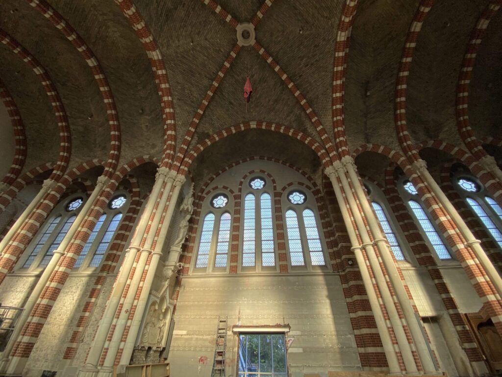 Interieur van een hoge, historische kerk met gotische ribgewelven, rood-witte bakstenen bogen en hoge glas-in-loodramen. Onderaan is een deuropening zichtbaar met een ladder ervoor, wat wijst op renovatie- of inspectiewerkzaamheden.