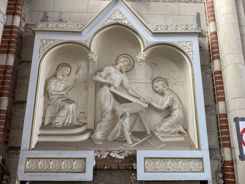Detail van een reliëfsculptuur in een kerkinterieur, met religieuze figuren in een nis onder gotische bogen. Onderin is schade zichtbaar in het metselwerk, wat wijst op restauratie- of herstelwerkzaamheden.