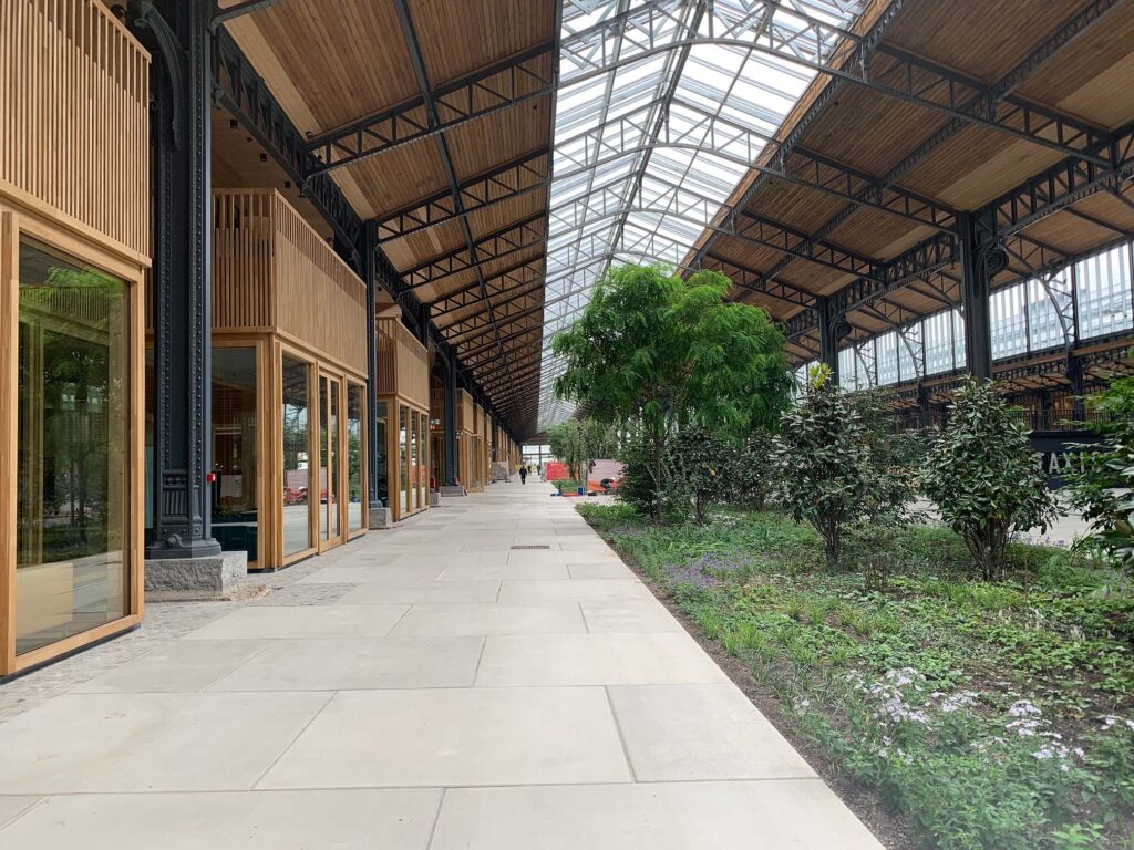 Overdekte binnenruimte in een gerenoveerd industrieel gebouw met glazen dak, houten gevels en groene beplanting langs een brede wandelgang.