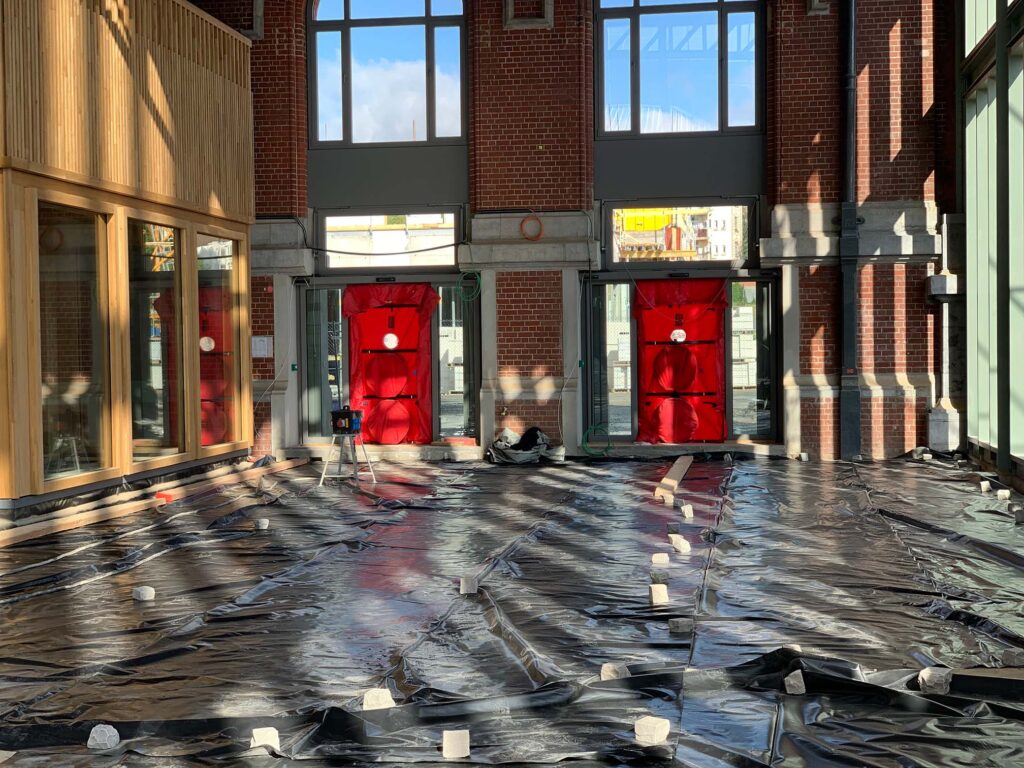 Blowerdoor testopstellingen in een grote hal van een gebouw in renovatie, met afgedekte vloer en meerdere deuropeningen voorzien van meetapparatuur voor luchtdichtheidsmetingen.