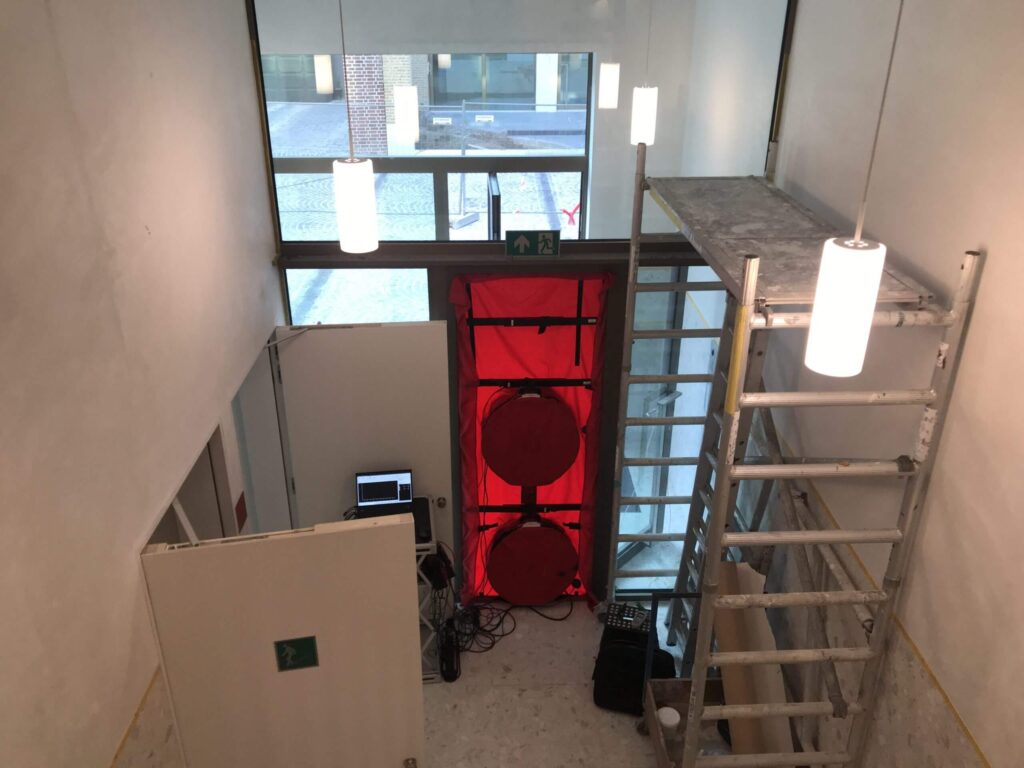 Blowerdoor testopstelling met ventilatoren in een entreehal, opgesteld in een deuropening met meetapparatuur en laptop voor luchtdichtheidsmeting.