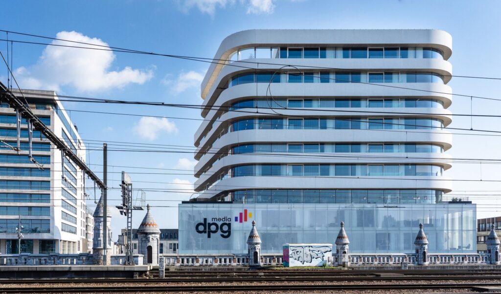 Modern kantoorgebouw van DPG Media met afgeronde verdiepingen en glazen gevel, gelegen langs spoorlijnen in een stedelijke omgeving.