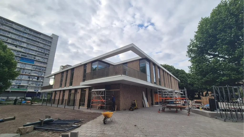 Nieuwbouw schoolgebouw met bakstenen gevel en overstekende verdieping, gefotografeerd tijdens de afbouwfase met steigers en bouwmaterialen op de voorgrond.