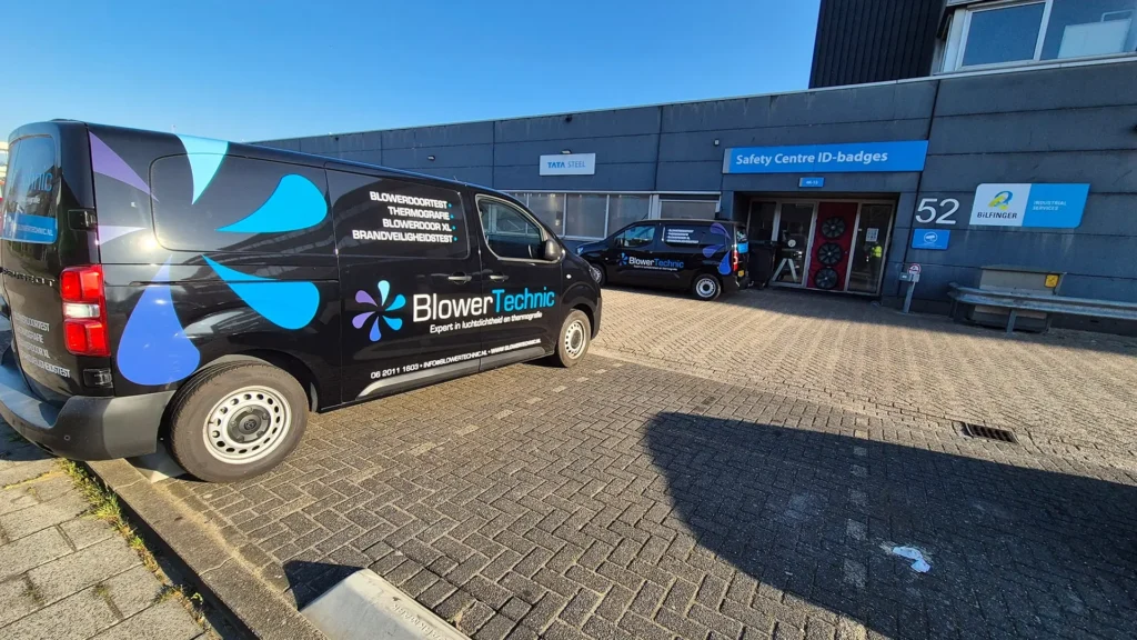 Bedrijfswagens van BlowerTechnic geparkeerd bij een bedrijfslocatie, waar een blowerdoor testopstelling zichtbaar is bij de ingang voor luchtdichtheidsmetingen.
