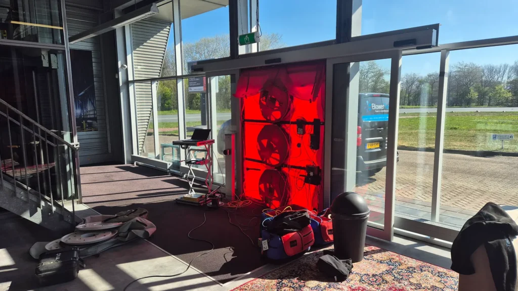 Blowerdoor testopstelling in een kantoorentree, met drie ventilatoren in een rode doekconstructie in de deuropening en meetapparatuur klaar voor luchtdichtheidsmeting.