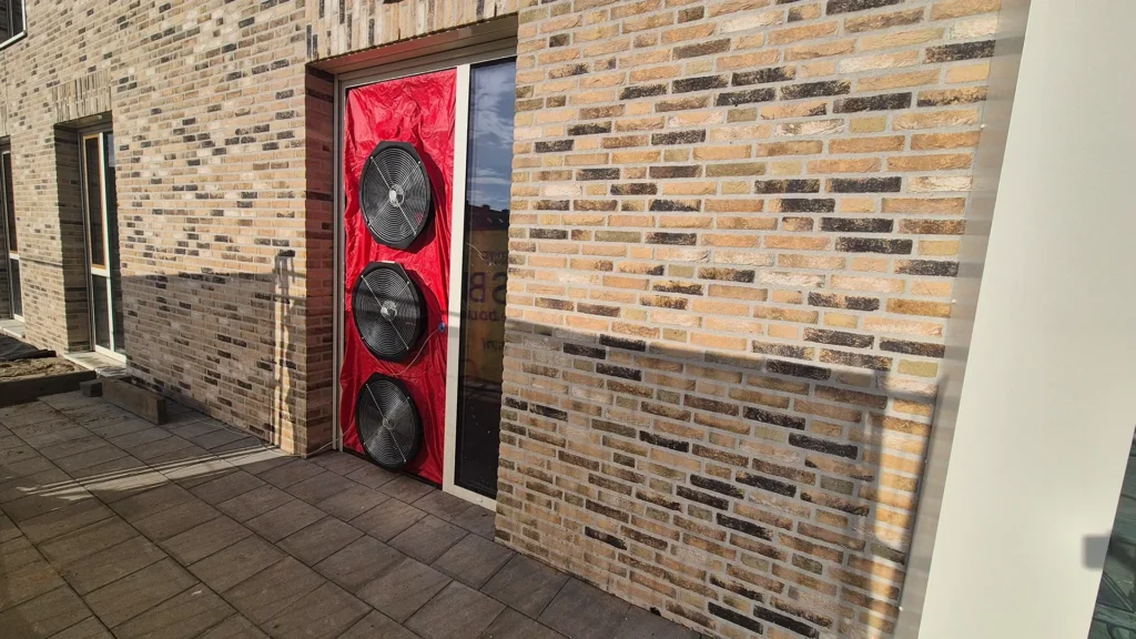 Blowerdoortestopstelling in de deuropening van een nieuwbouwcomplex, met drie ventilatoren in een rode afdichtingsdoek om de luchtdichtheid van het gebouw te meten.