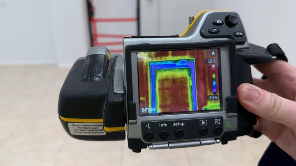 Thermografische camera die een deuropening in beeld brengt met temperatuurverschillen, gebruikt voor het opsporen van luchtlekken en warmteverlies.